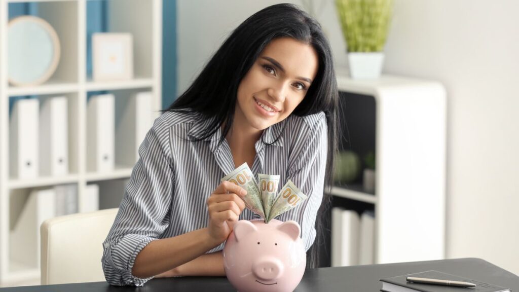 Woman Putting Cash Money In A Pink Piggy Bank In An Office