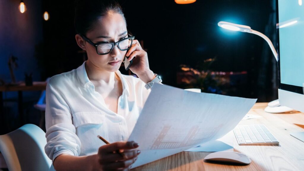 Serious Woman With Glasses Reading A Paper Report While Phone On Her Ear