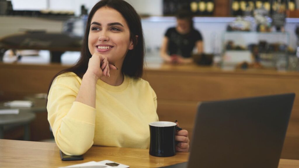 Lady Smiling Looking At The Left While Holding A Cup In Front Of A Laptop