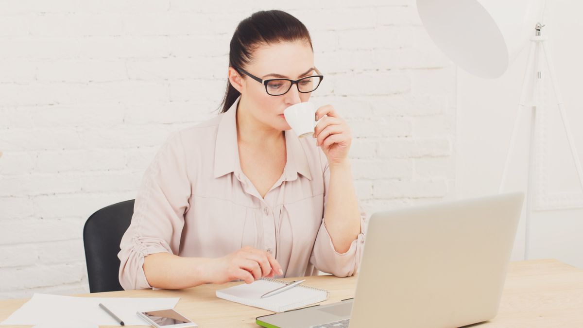 Woman With Glasses Sipping Coffee Cup While Reading On Laptop