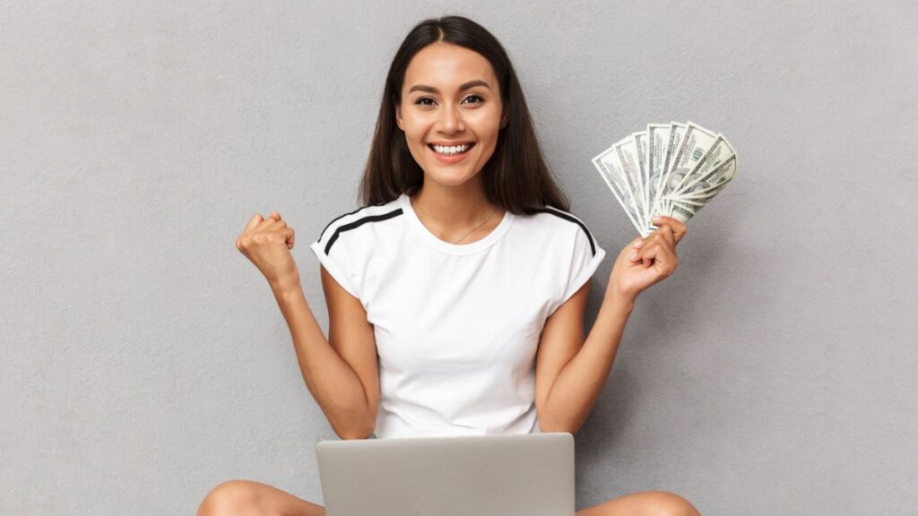 Happy Young Woman Sitting With Laptop On Her Lap Holding Cash Money