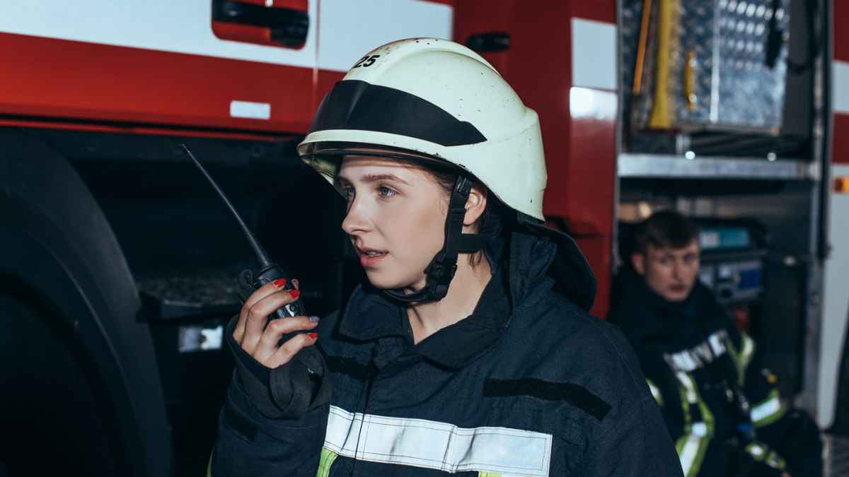 Female firefighter talking on portable radio set with colleague behind.