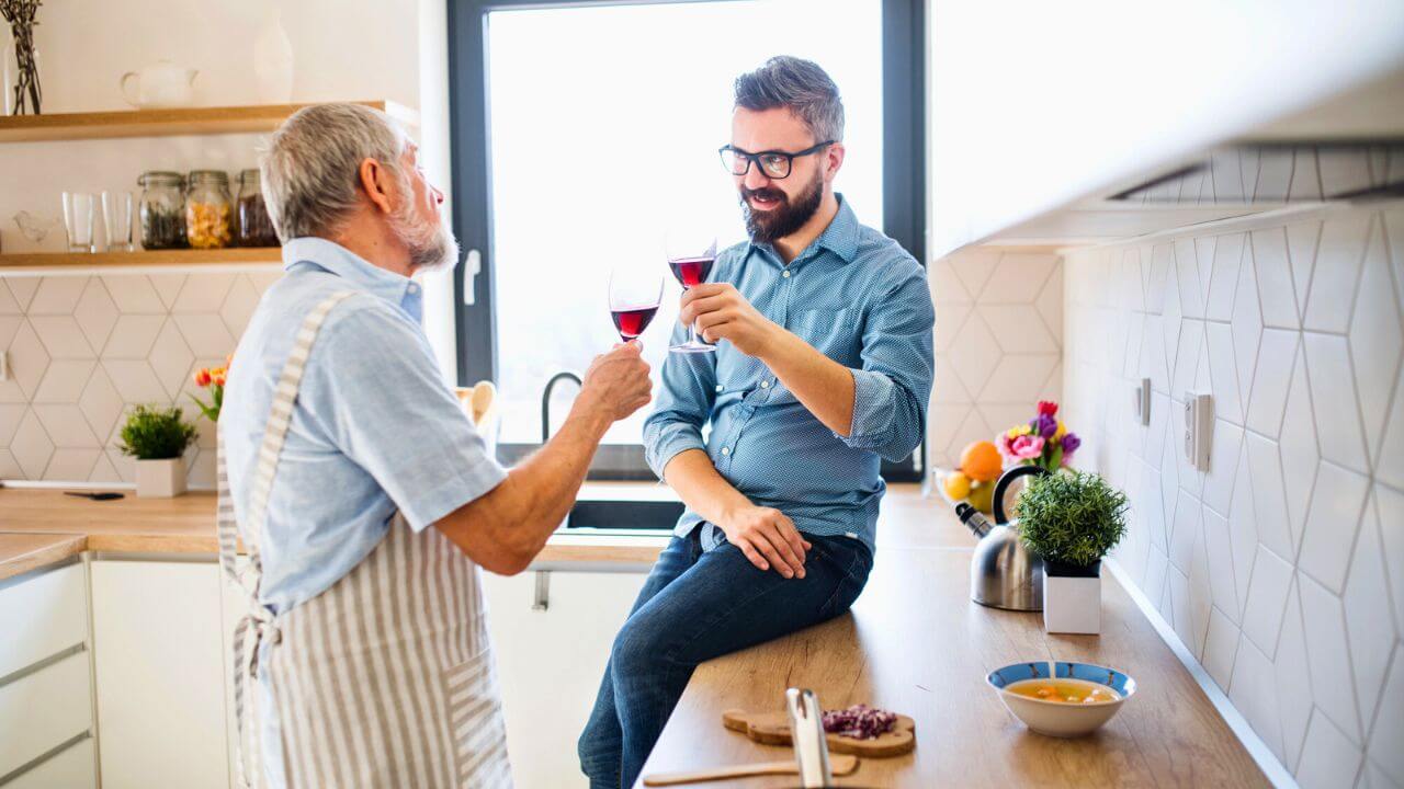 An Adult Hipster Son and Senior Father Indoors in Kitchen at Home, Drinking