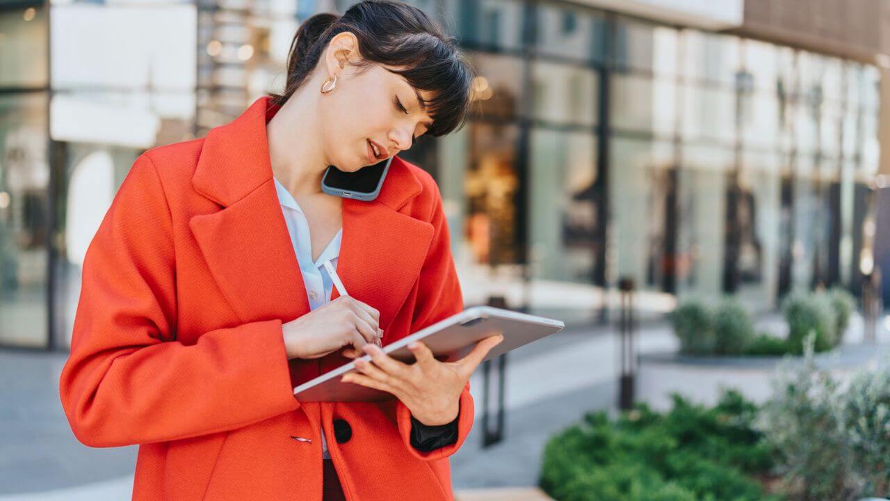 Businesswoman in Red Coat, Multitasking with a Tablet and Phone